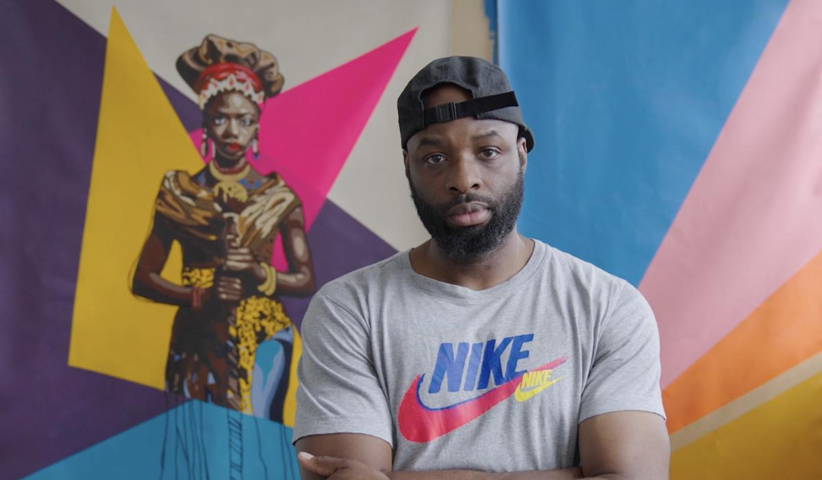 Artist Joseph Ijoyemi stands in front of a colourful painting depicting a queen against a backdrop of colourful triangles