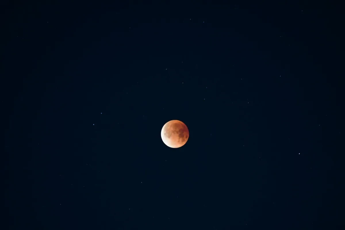 Photo of the Moon which is 90% illuminated in a rust red hue against a starry black sky