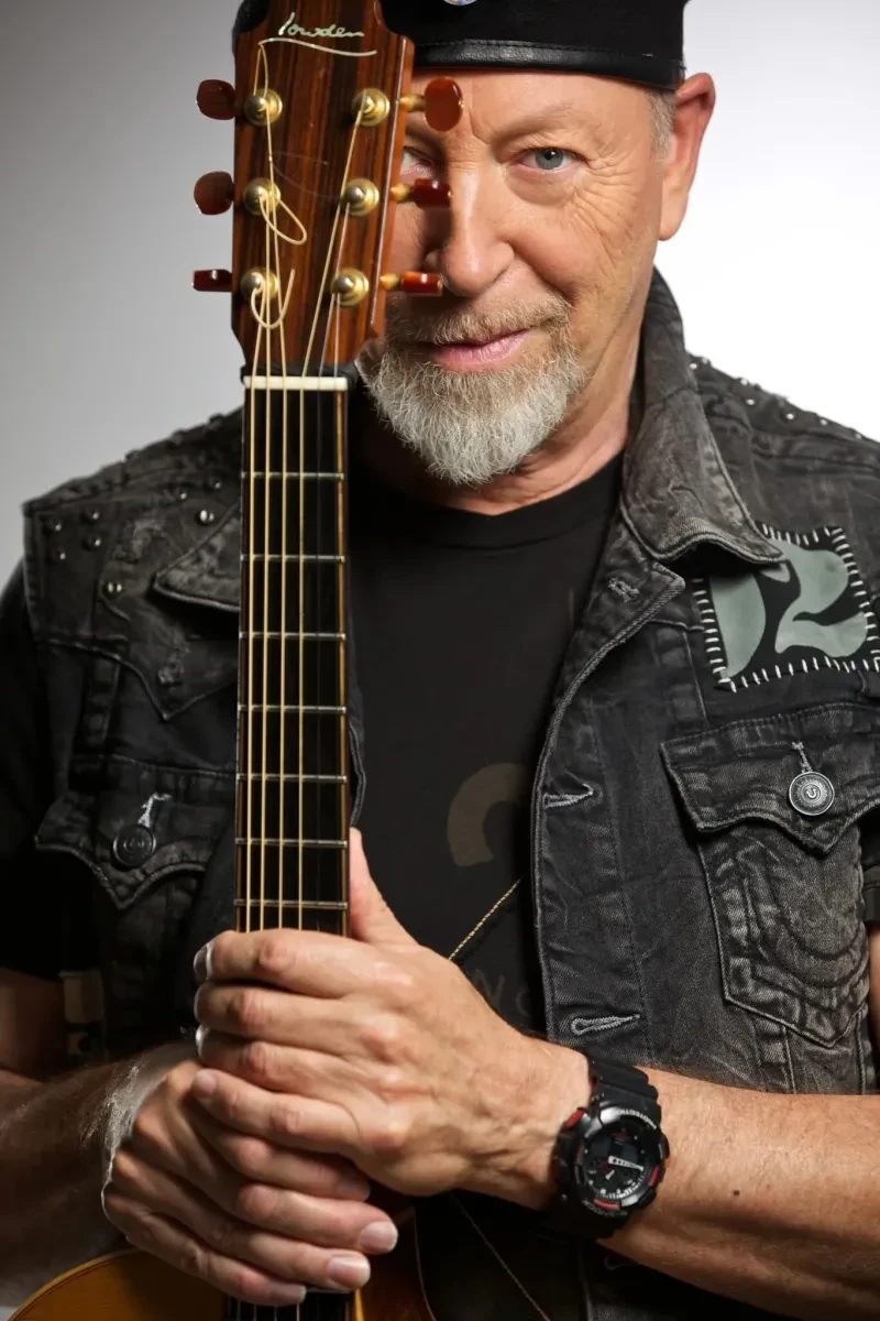 Photo of Richard Thompson who has a black hat and white goatee holding up a guitar in front of half of his face