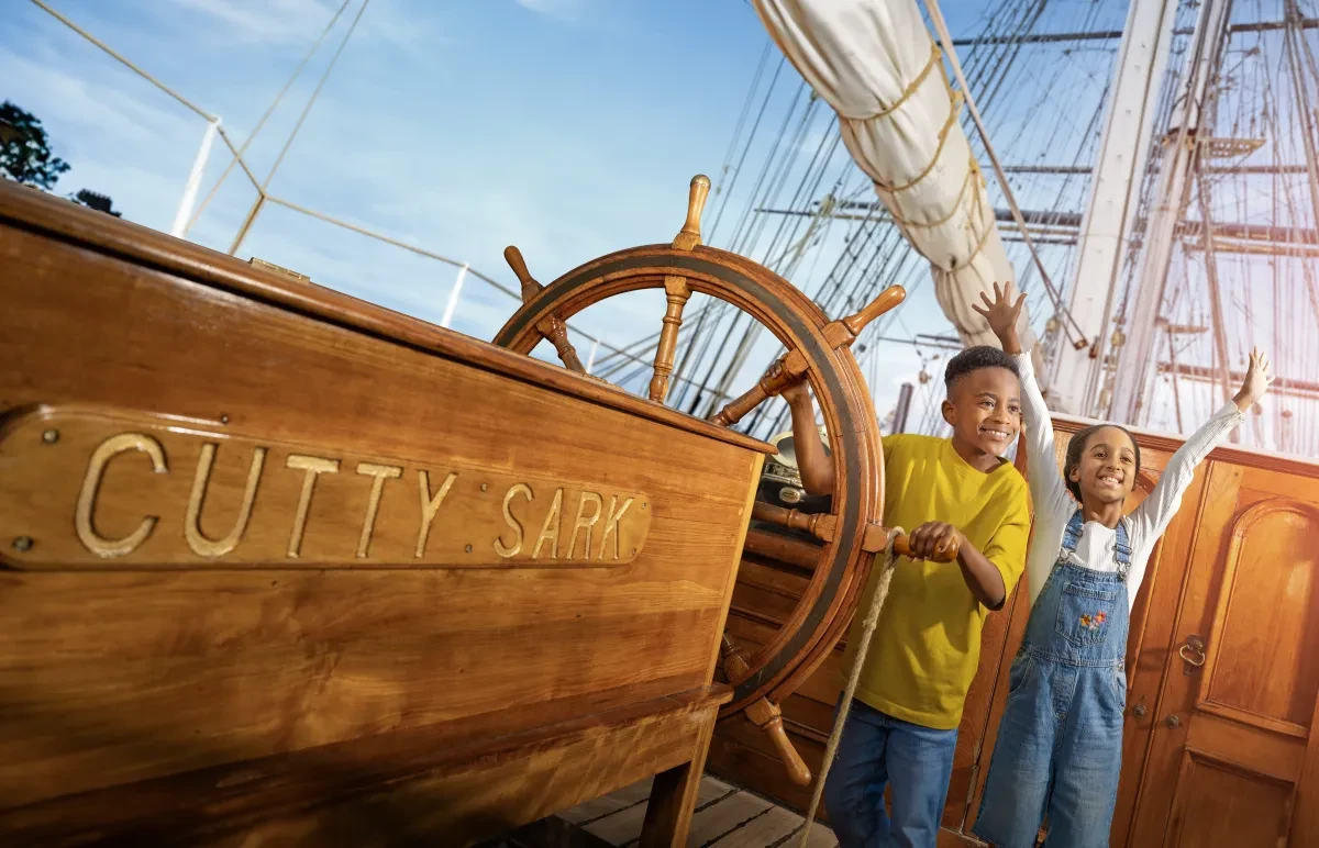 Photo of two children on board a historic sailing ship next to the ship's wheel looking happy and excited