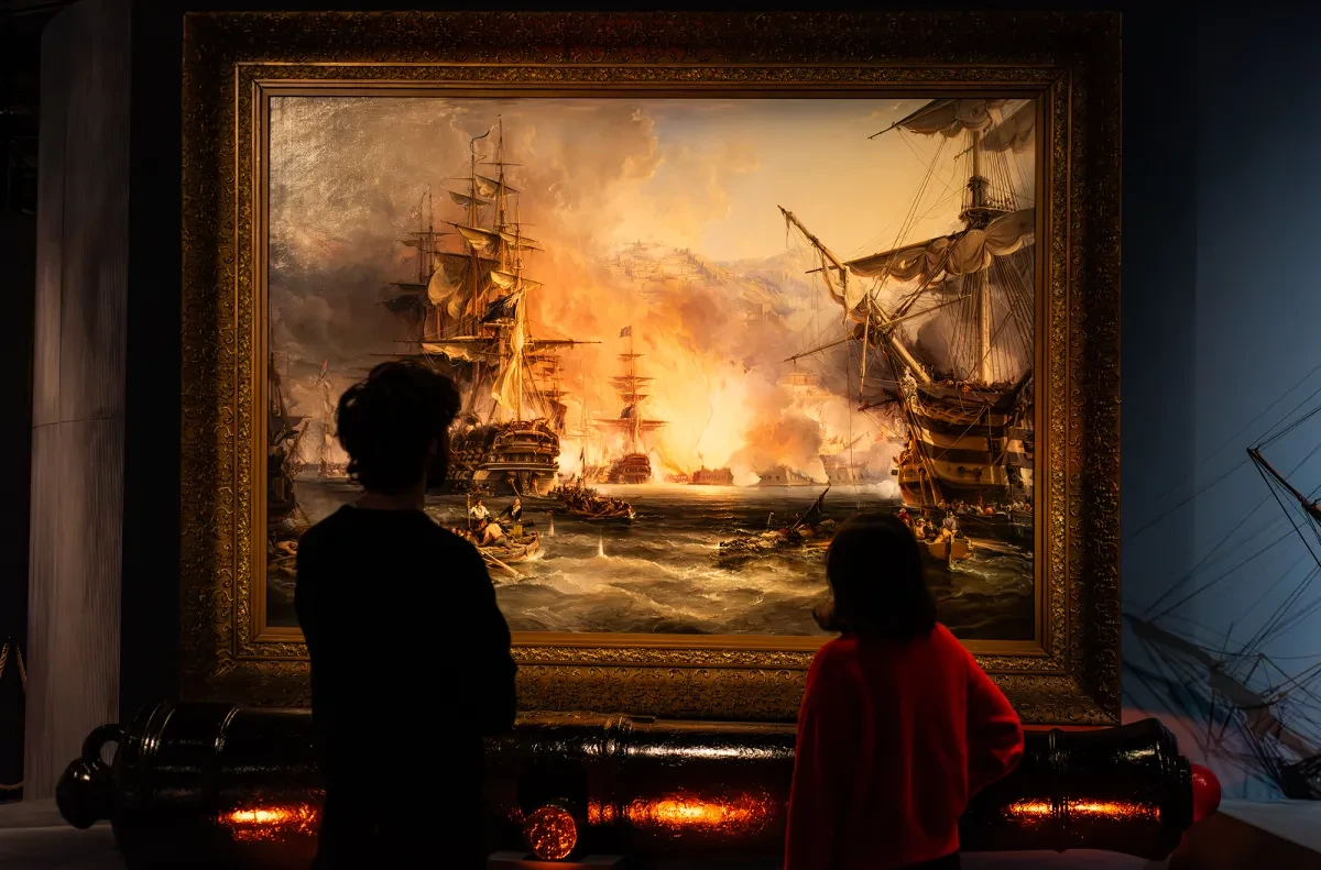 Two people look up at a painting depicting the Bombardment of Algiers in the Pirates exhibition at the National Maritime Museum