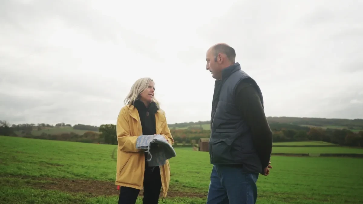 A still from the film Fabric of the Land: The Untold Yarn, showing artist Ella Doran examining a piece of textile design with a farmer in a field