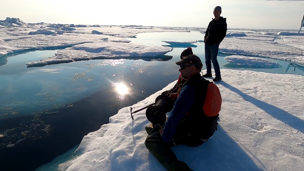 Two figures kneel on the ice and look out at a landscape of broken ice and seawater. Another figure is standing behind them, silhouetted against the Sun