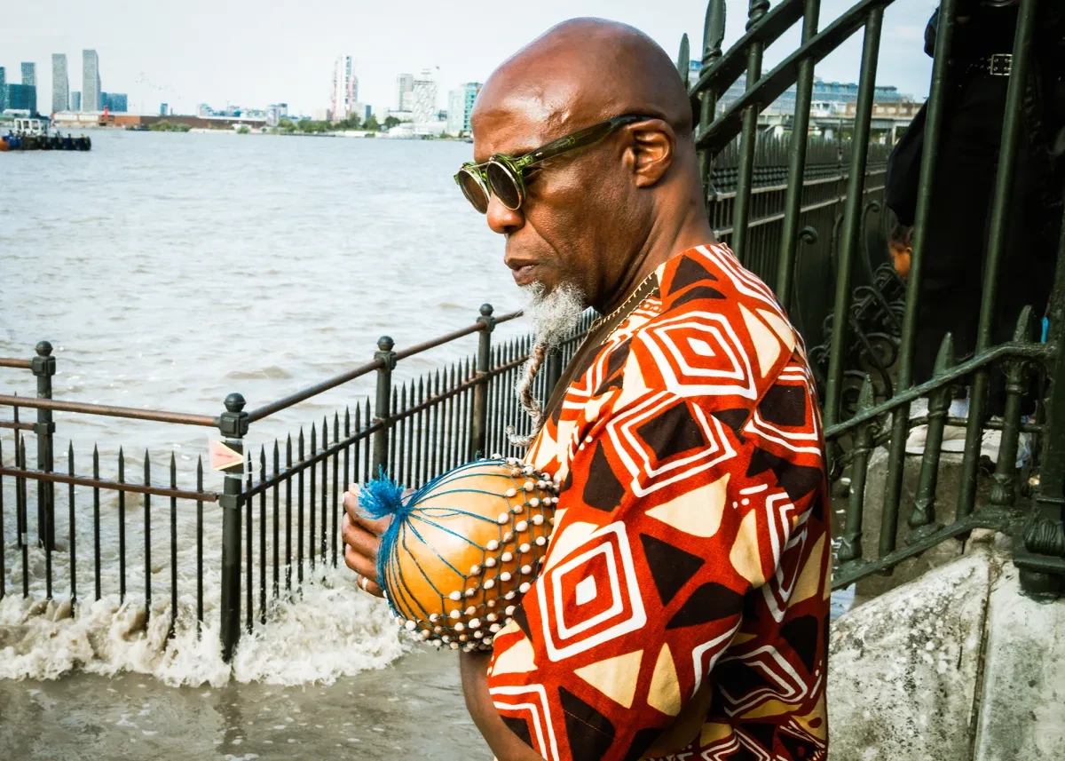 A Black man wearing green sunglasses and a geometric patterned top stands contemplatively by the River Thames