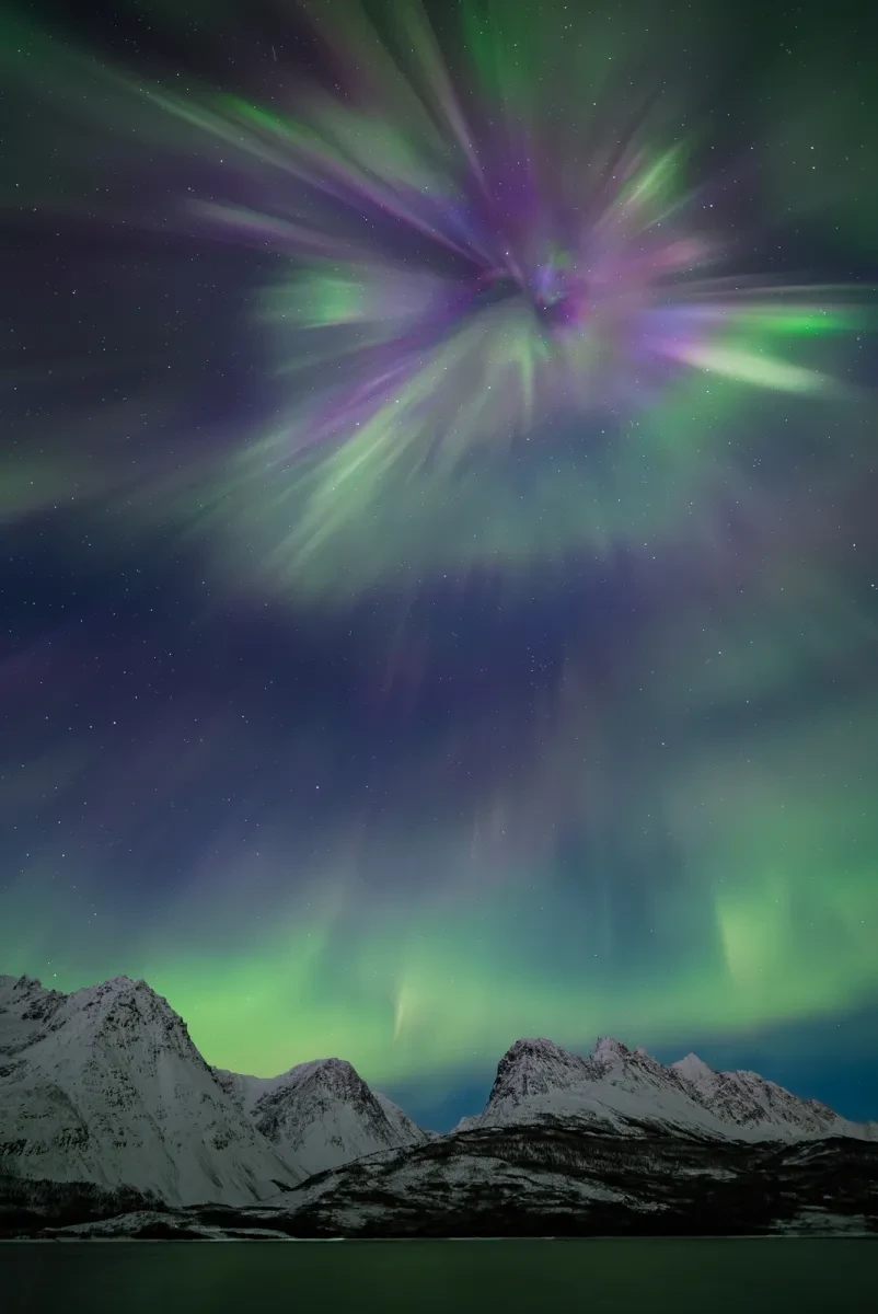 Photo showing icy snowy mountain landscape with vast sky above filled with green and purple aurorae, at the top is a large firework-shape aurora in green and purple