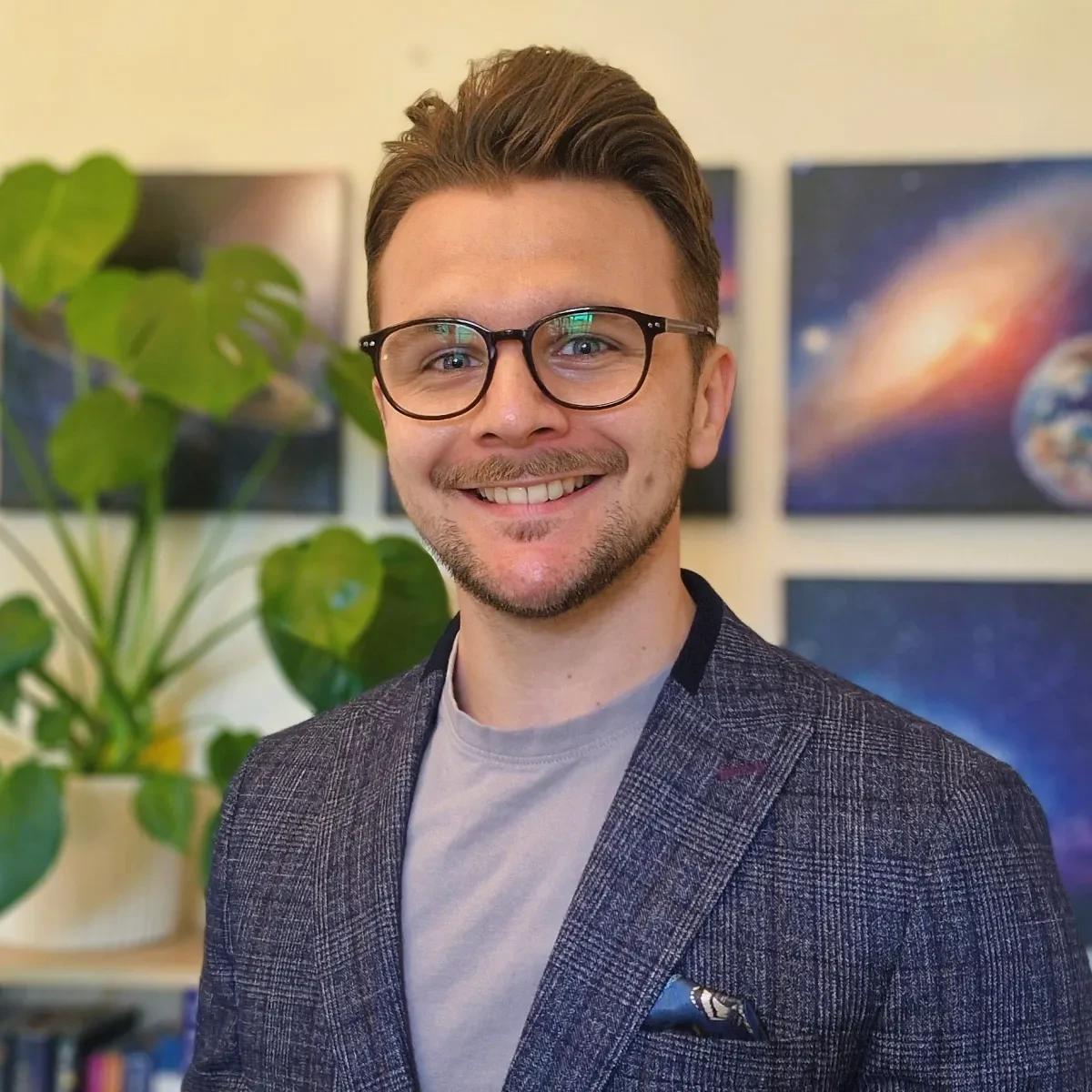 Astronomer Jake Foster smiling brightly. He has glasses with dark frames, light stubble and is wearing a chequered navy jacket and light grey t-shirt