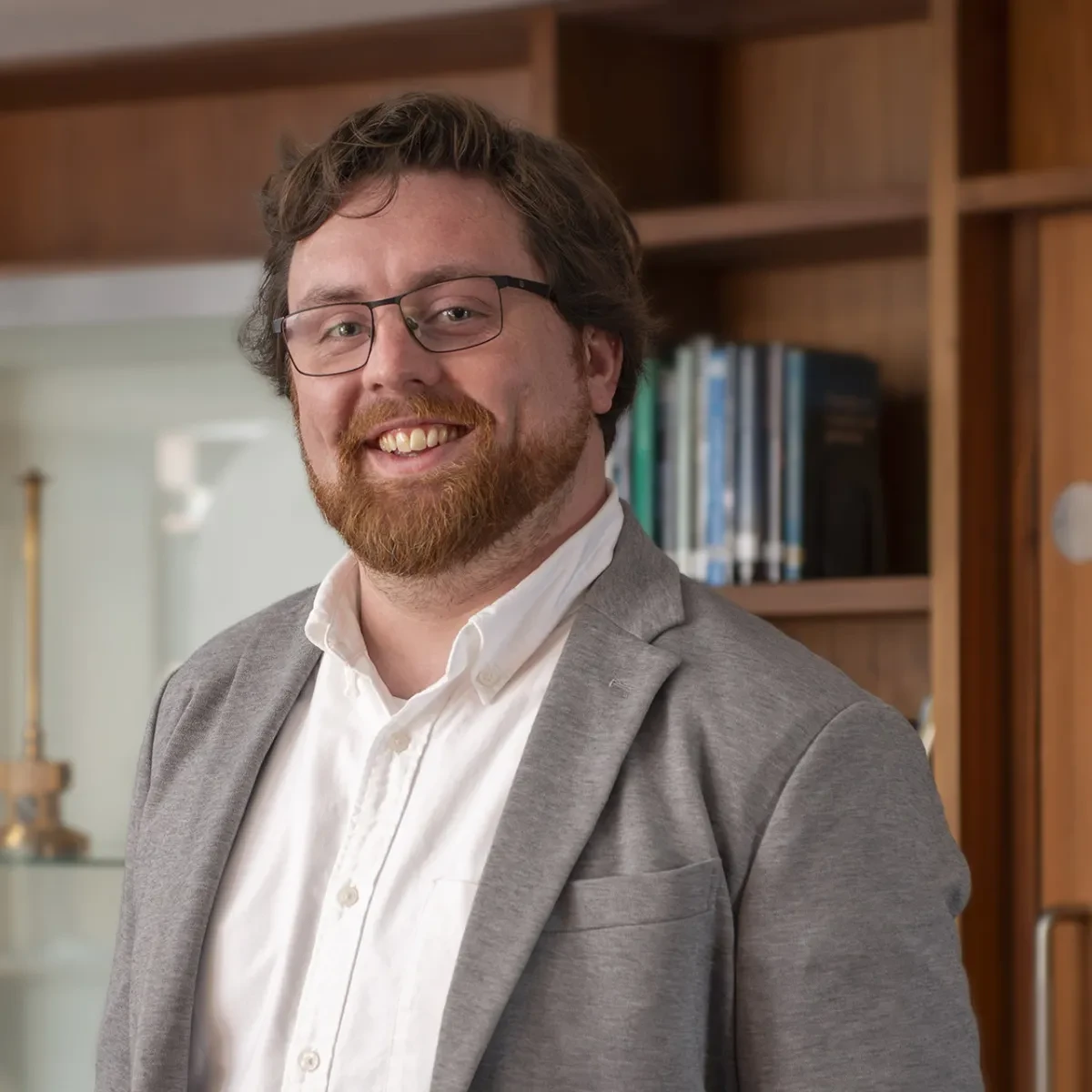 Astronomer Dr Ed Bloomer looking happy and relaxed. He has dark curly hair and a trimmed bear, and is wearing glasses and a light grey suit