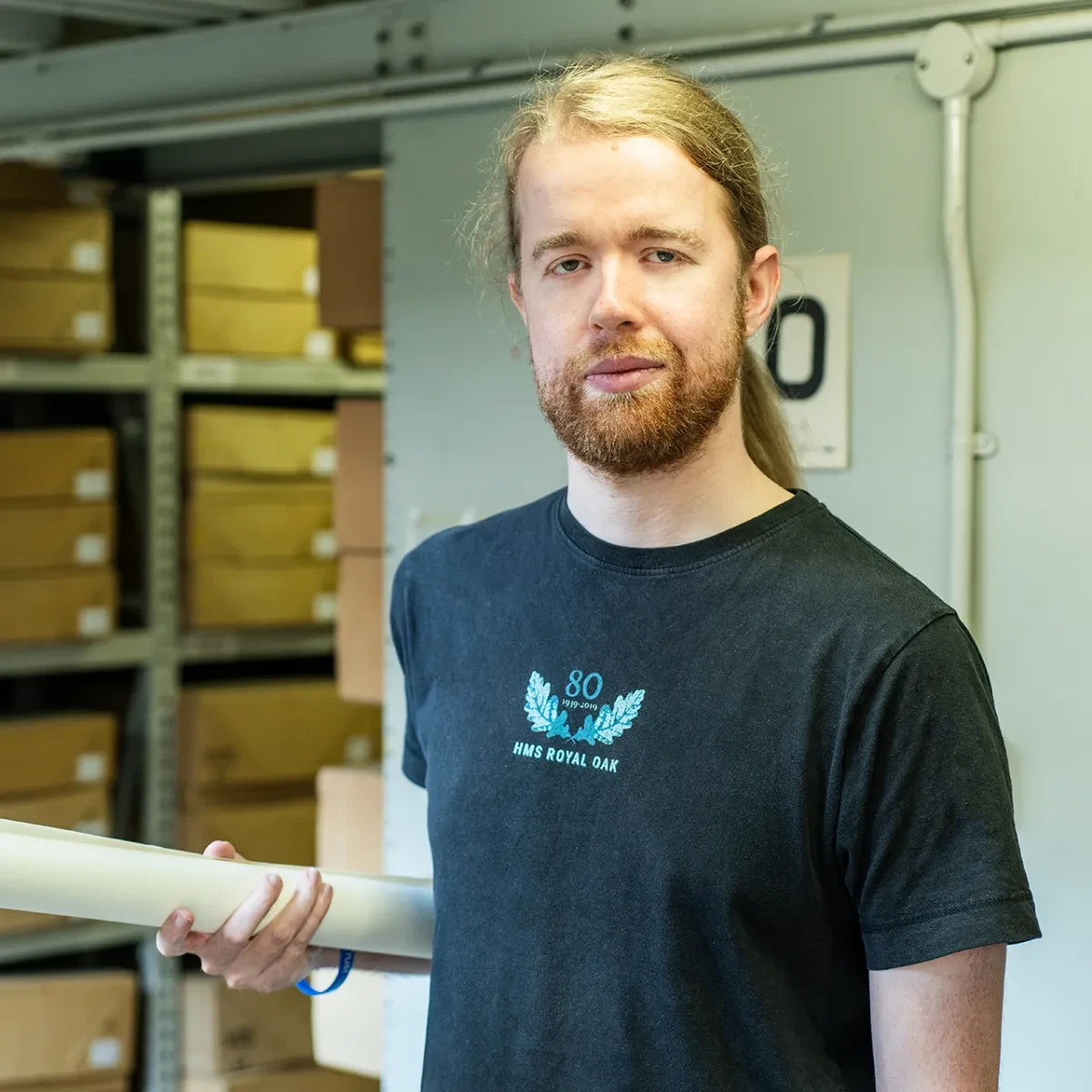 Headshot of curator Alex Grover. He has a light brown beard and hair, and is standing with a roll of paper under one arm