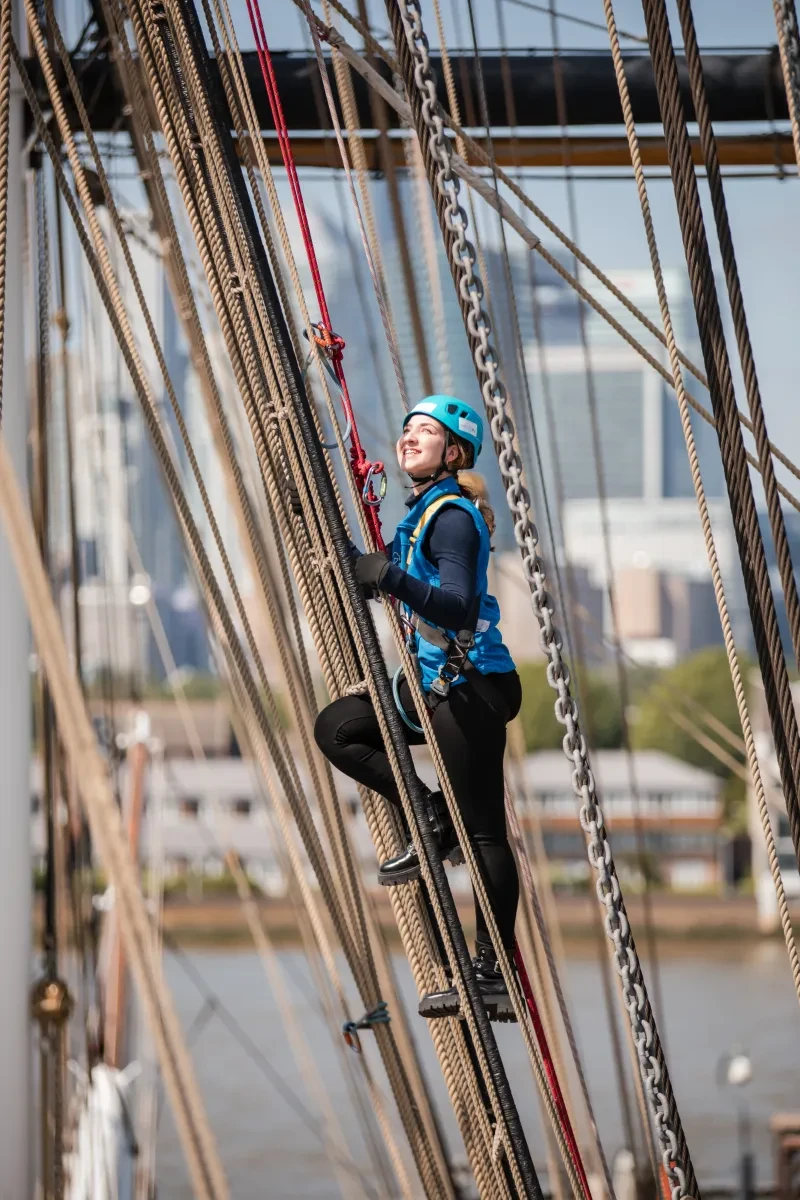 A woman climbs the rigging of tall ship Cutty Sark. She is looking up and smiling as she takes part in the experience