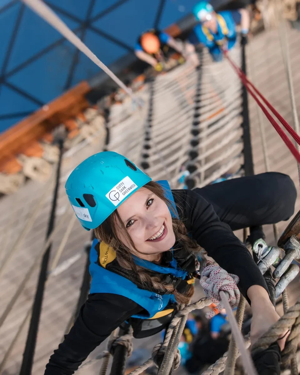 A woman wearing a blue helmet and harness looks up as she holds on to the rigging of a ship. Far below her the deck can be seen