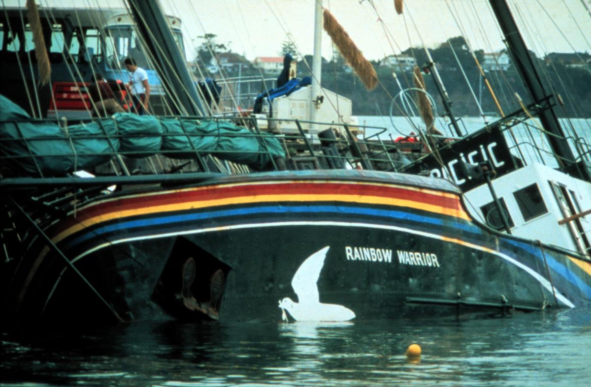 The ship Rainbow Warrior partially sunk in Auckland harbour. A rainbow band and the white outline of a dove are visible above the waterline on the ship's hull