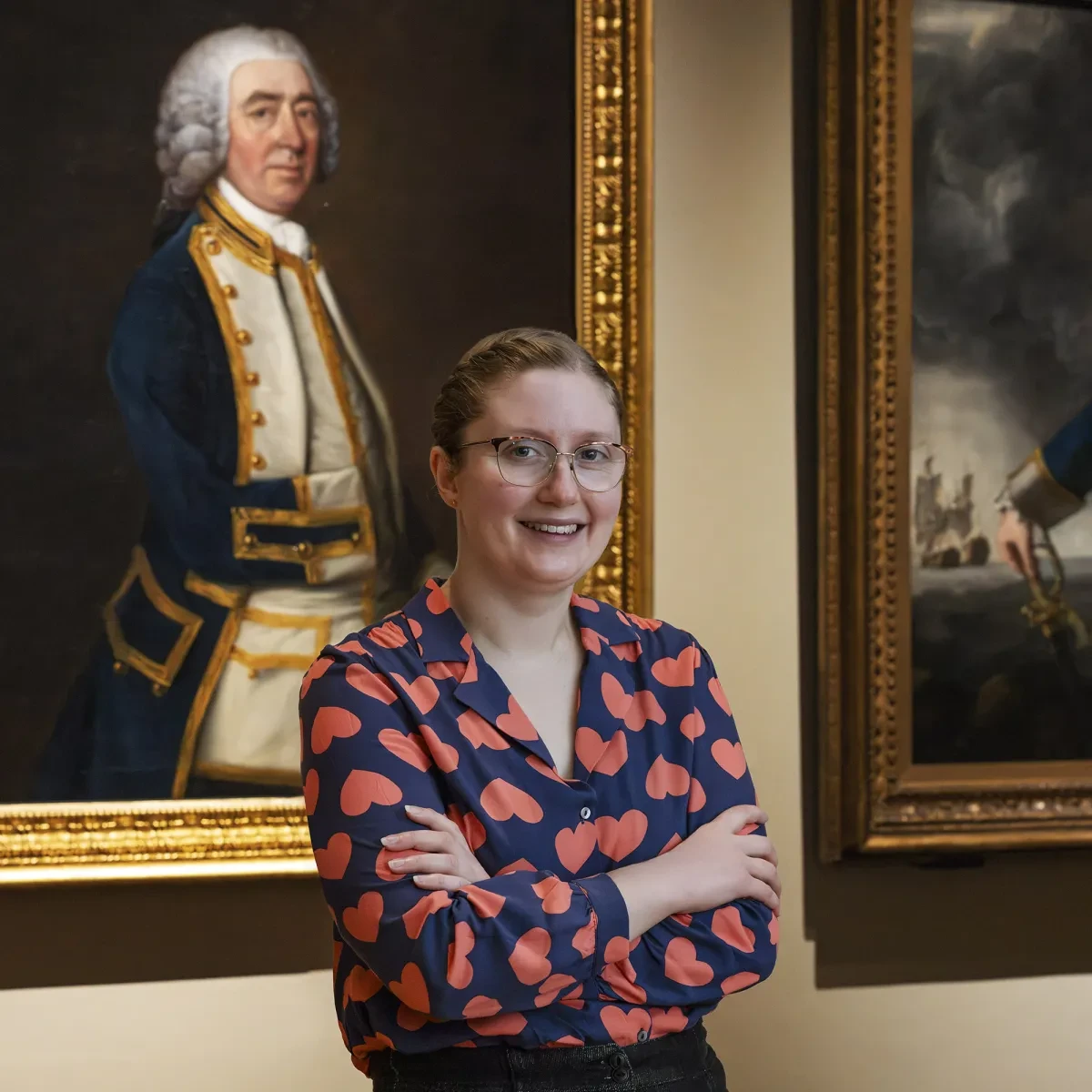 Curator Katherine Gazzard standing in front of a grand portrait of a naval officer. Katherine is wearing glasses and a navy blouse decorated with red hearts