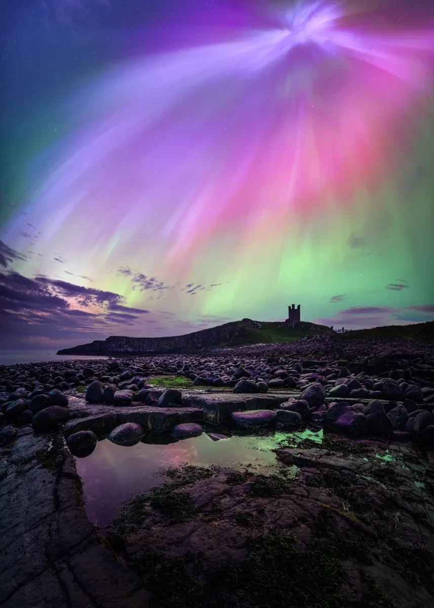 A dark view looking across a bay, with a castle turret in the distance. Above the scene the aurora appears as a cascade of purple, pink and green light