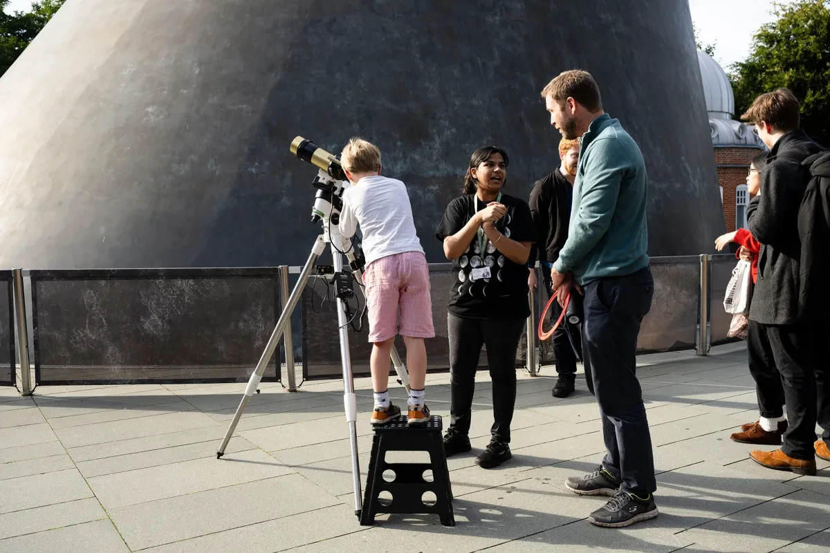 A boy stands on a box to look through a telescope outside, as an astronomy guide and adult look on