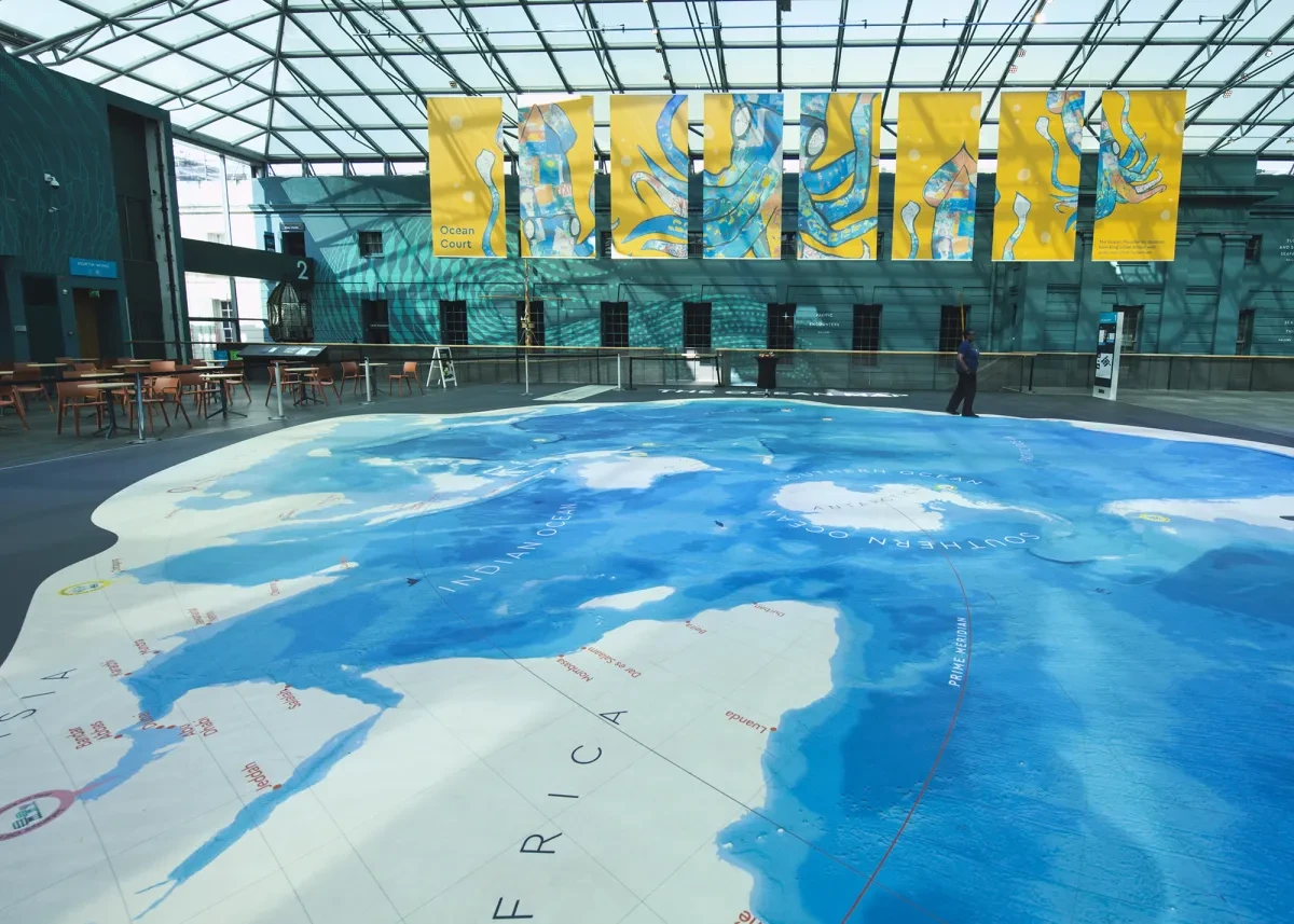 Inside Ocean Court at the National Maritime Museum. Colourful banners hang from the glass ceiling, while a large floor map of the ocean is visible in the foreground