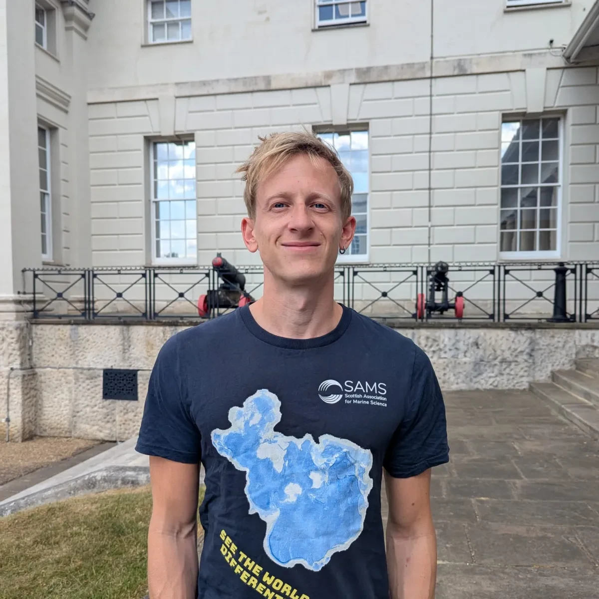 Curator Tim May stands in front of the National Maritime Museum. He is wearing a t-shirt featuring a type of map known as a 'Spilhaus projection'