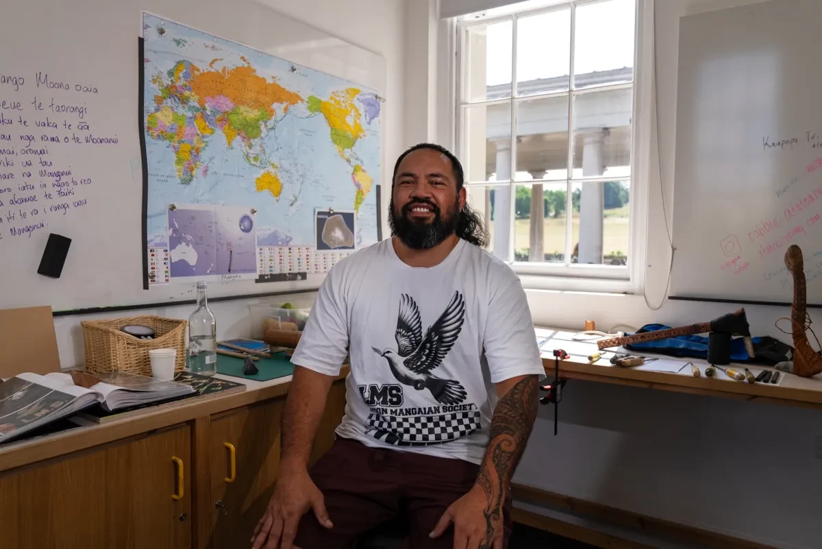 A photo of Robbie Atatoa, sitting on a stool in the artist studio wearing a white t-shirt with a bird and traditional Mangaian patterns below plus a title saying London Mangaian Society. There is a world map in the background and Robbie's carving tools on a work bench behind him