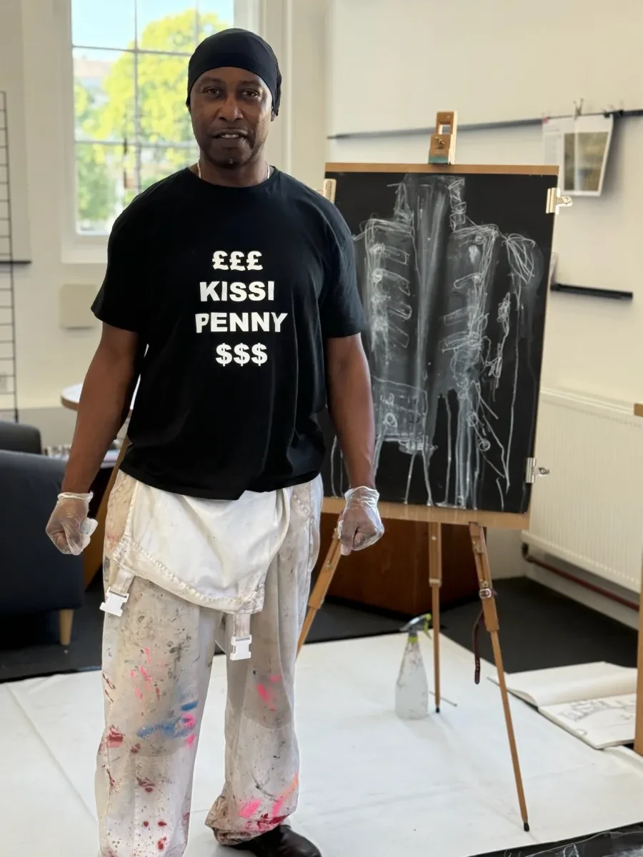 Evewright is a Black male adult, wearing a black durag and black tshirt with printed text. He wears stained overalls tied at his waist, and is smiling in front of an art piece depicting a sketched naval uniform on a black background.