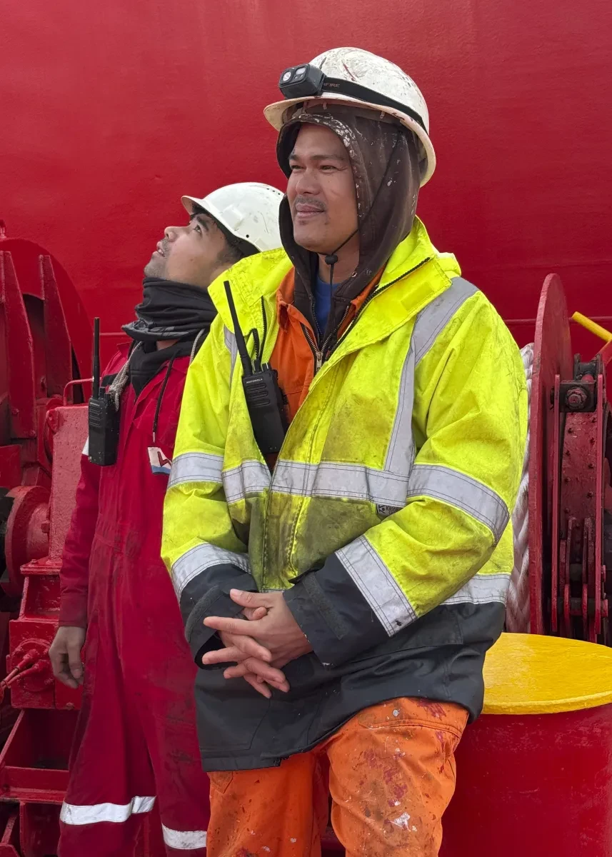 Two seafarers stand on deck of a large container ship. One in front wears a yellow hi-vis jacket over orange overalls and a white hard hat with a head torch strapped to it. The man behind is looking up at something and wearing a red boiler suit