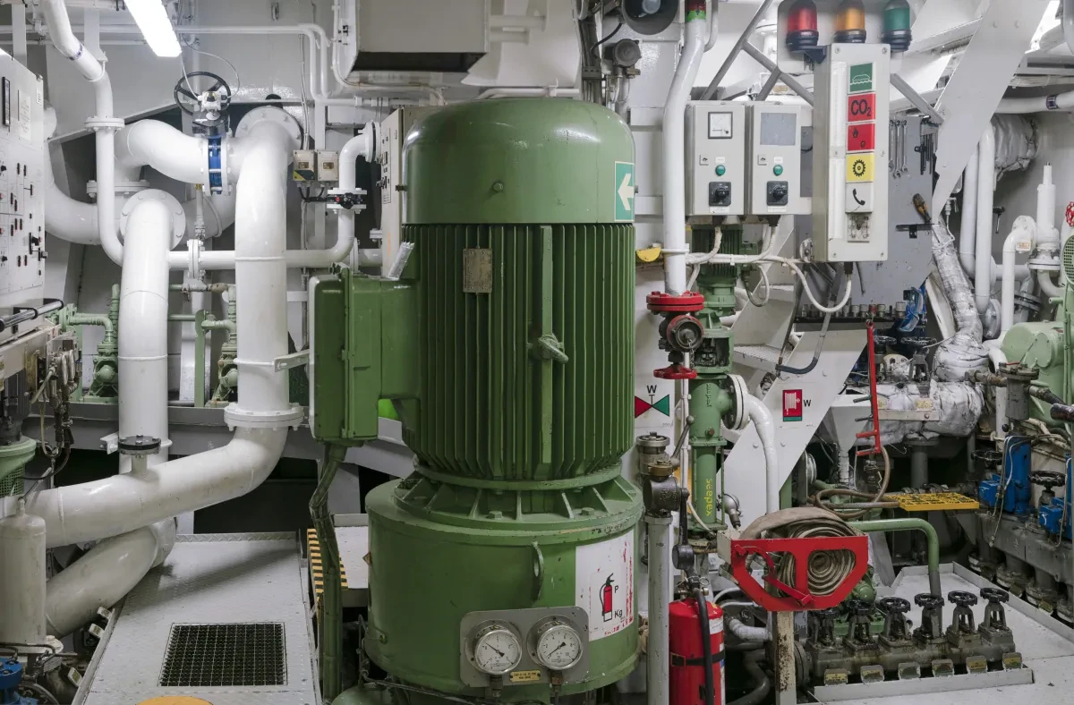 Interior or a container ship engine room. A large green cylinder is in the centre of the image, with numerous white pipes and switches surrounding it