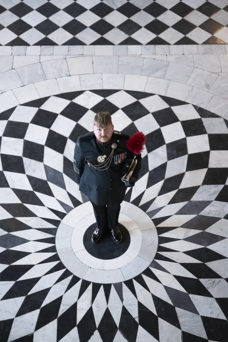 Colour photo of a white man in uniform, standing in the great hall which is a B&W tiled floor. Photo taken from above.