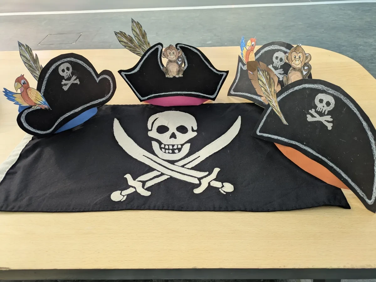 Four paper pirates hats, decorated with paper parrots and monkeys sit above a black and white Jolly Roger flag
