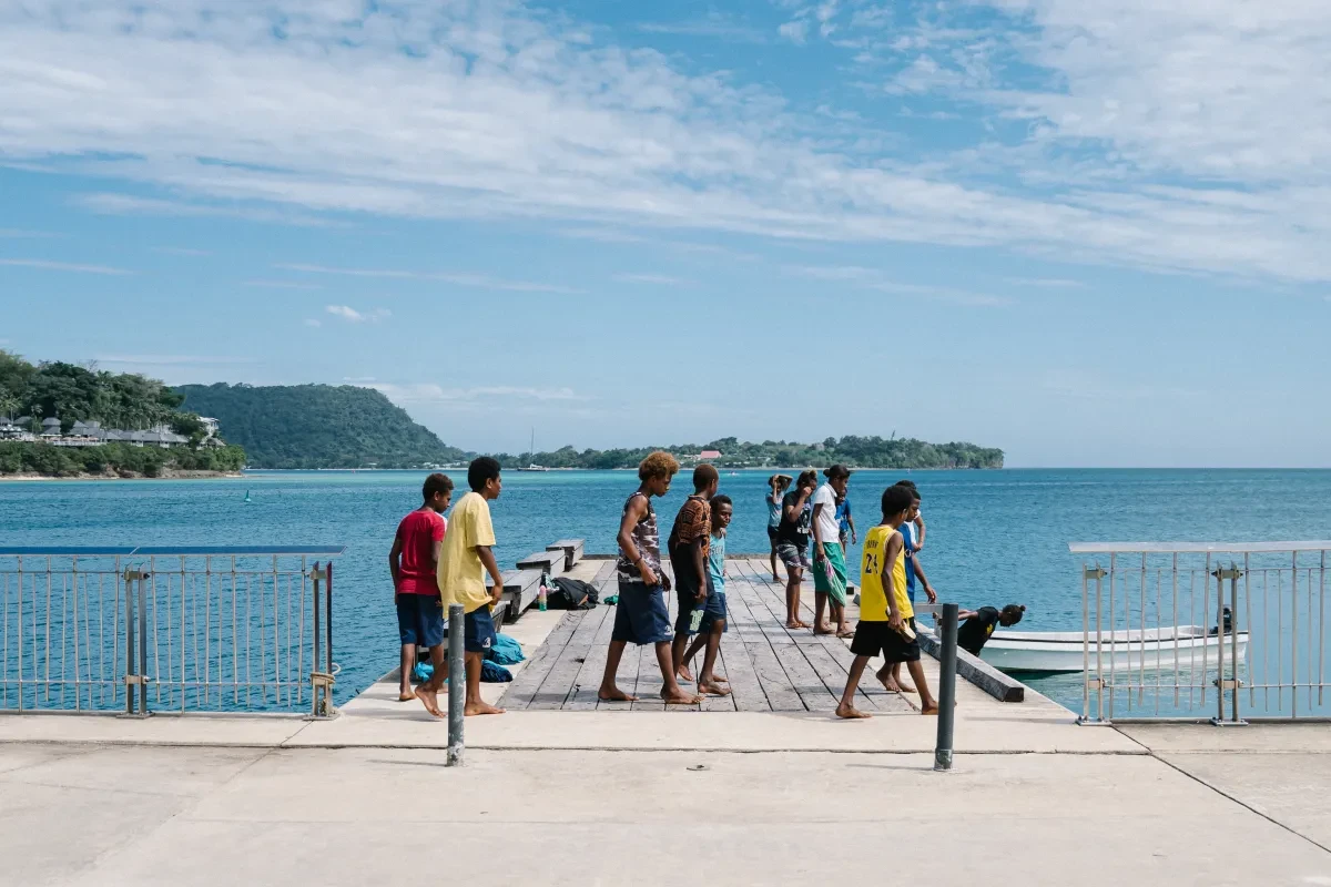 A group of young kids in colourful shorts and t-shirts play by a wharf jutting out into a sparkling blue sea