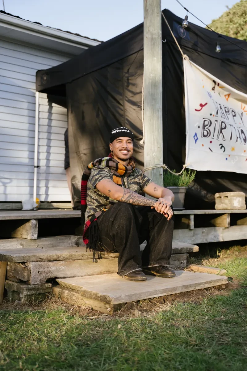 A smiling young man wears trousers and a shirt in muted colours, and sits on the step in front of a white, wooden building