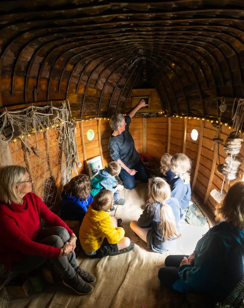 A group of children sit inside the upturned hull of a wooden boat, which has been converted into a miniature museum