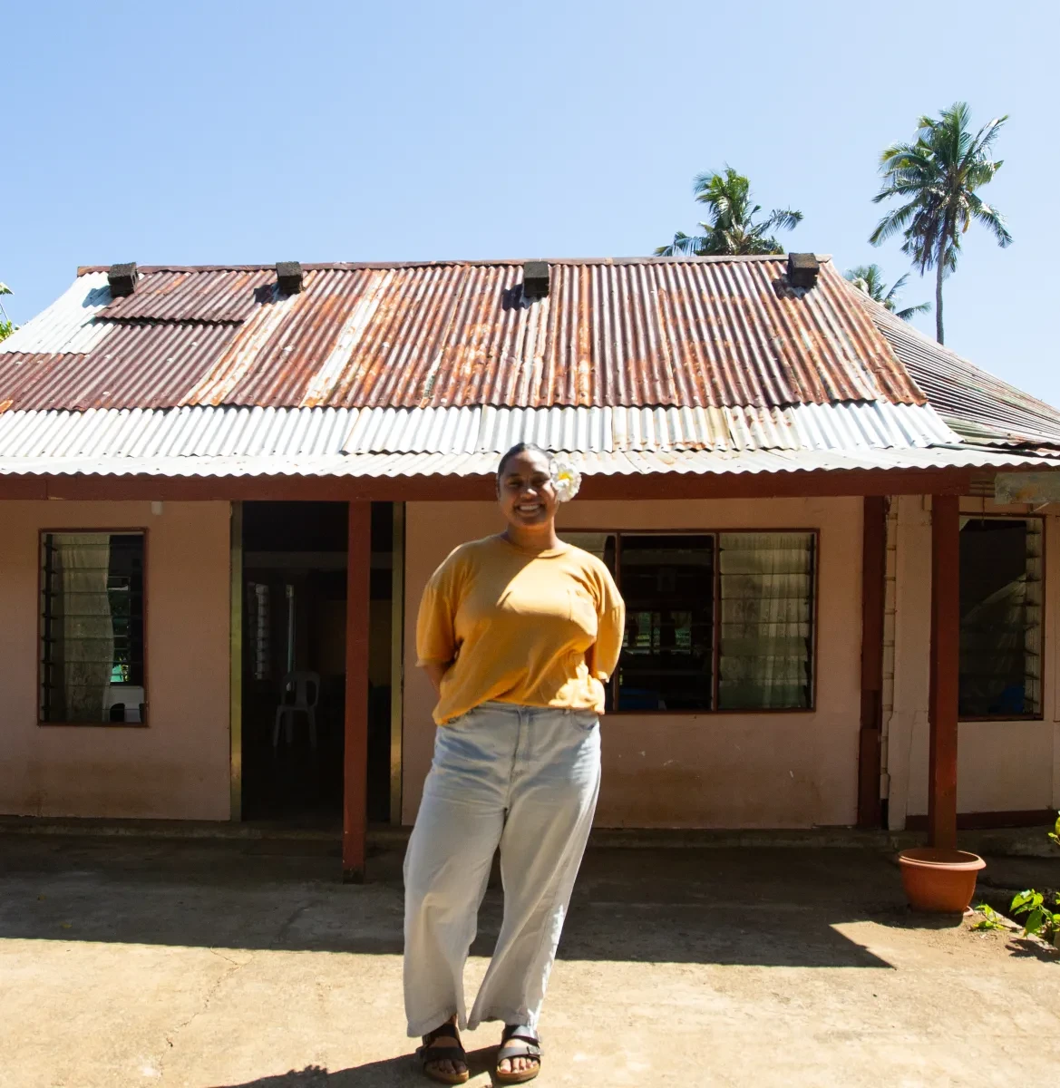 A woman in a relaxed yellow tshirt, beige trousers and sandals stands smiling in front of a bungalow with palm trees in the background