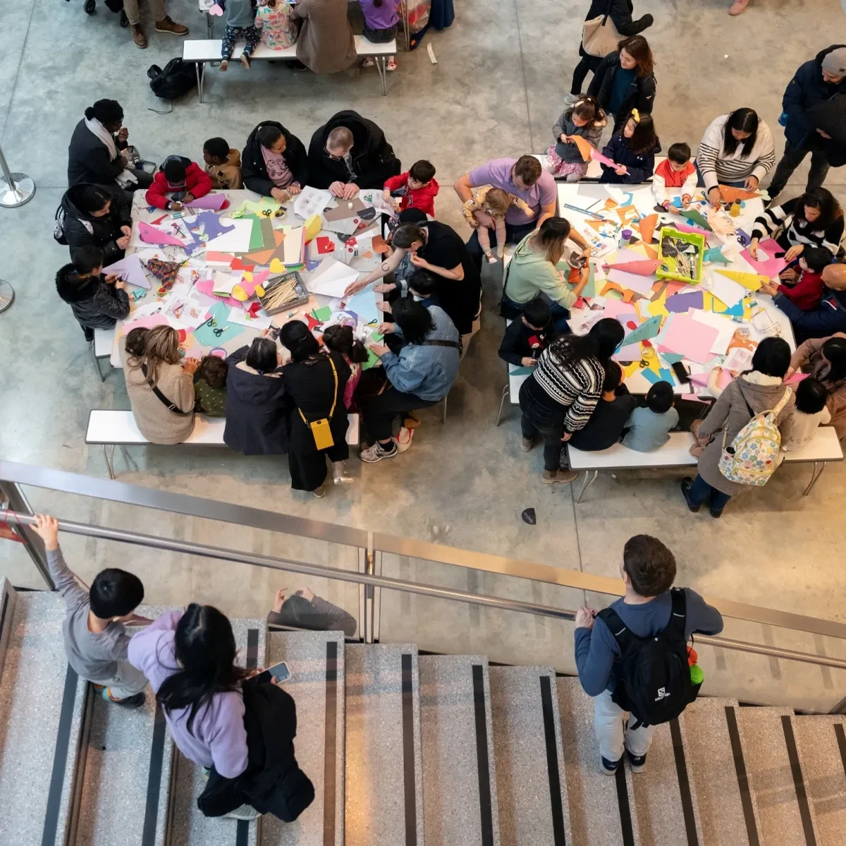 Overhead view of children and adults making art at a table with paper, scissors, and glue. Others watch nearby on a staircase