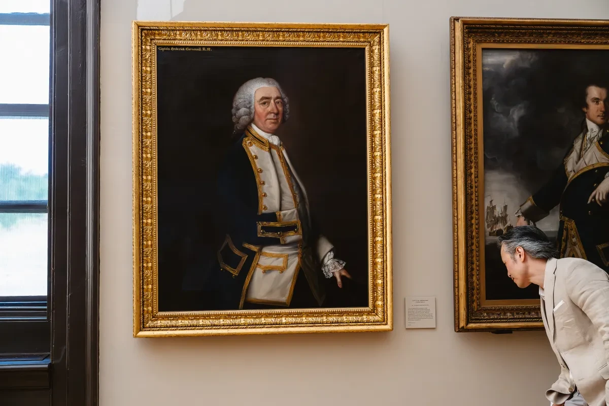 A man looks closely at the label next to an oil painting by Thomas Gainsborough of a man in a wig and historic naval attire in a gold frame in the Queen's House in Greenwich