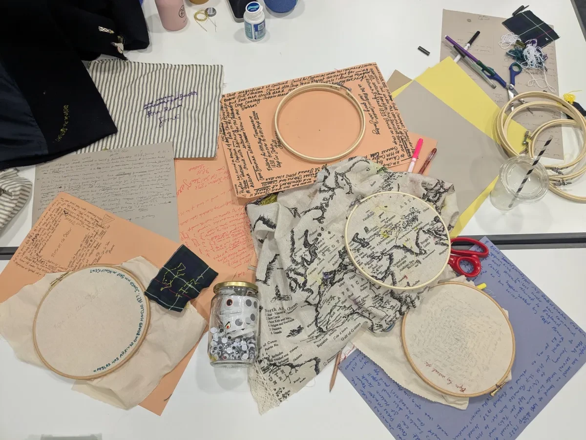 A mass of crafting materials: orange, purple and grey paper with writing going around the four edges, plus embroidery hoops with the start of embroidered texts, map-printed fabric and other designs, and scissors and pens. 
