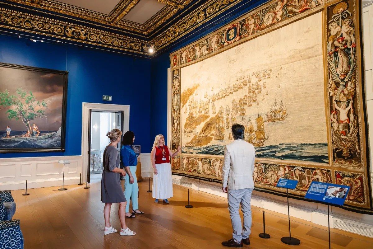 Three visitors in summer dress stand and admire the huge Solebay Tapestry in the Queen's House alongside an art curator