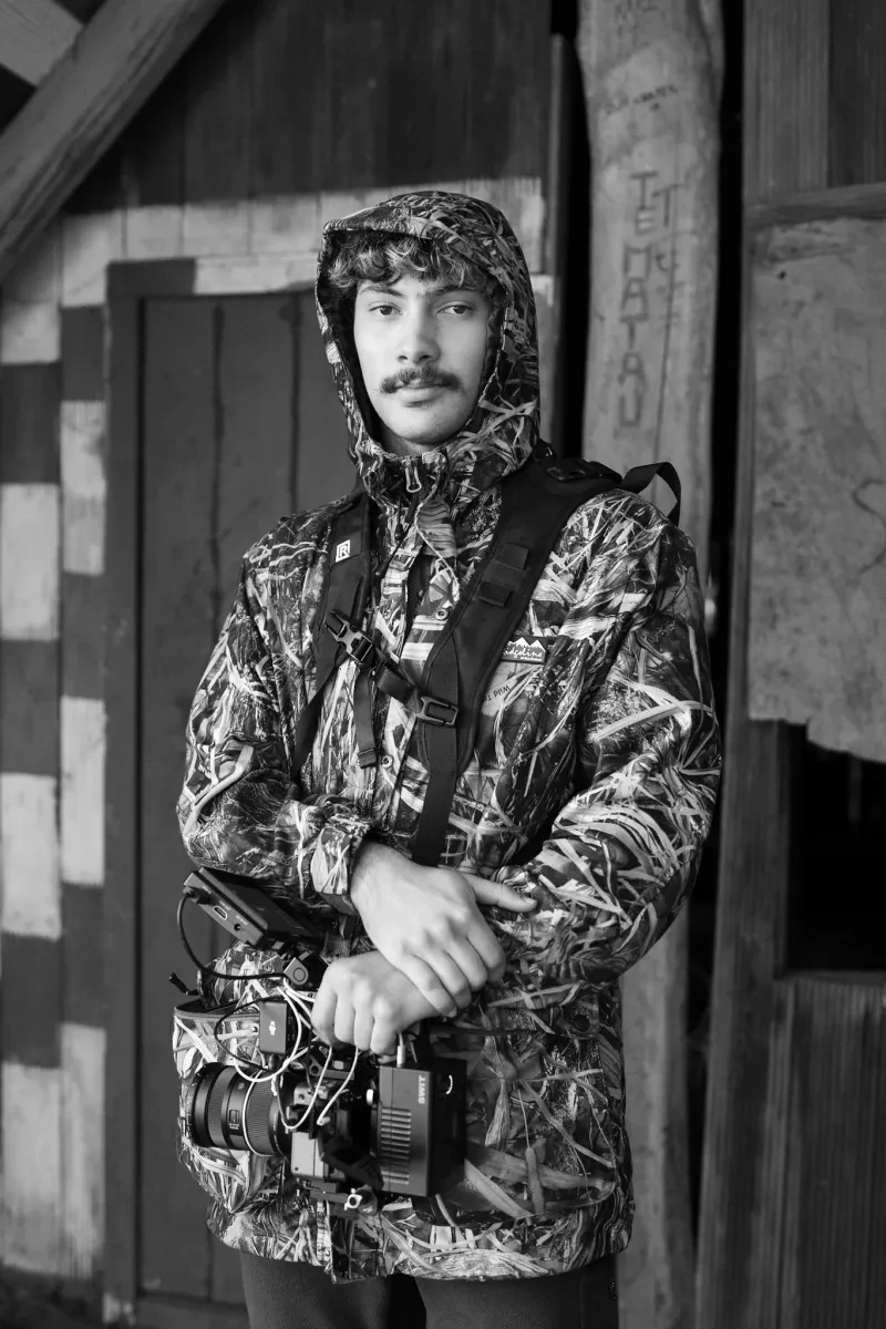 A black-and-white photo of a young man with a moustache wears a hooded coat and carries a camera strapped across his body