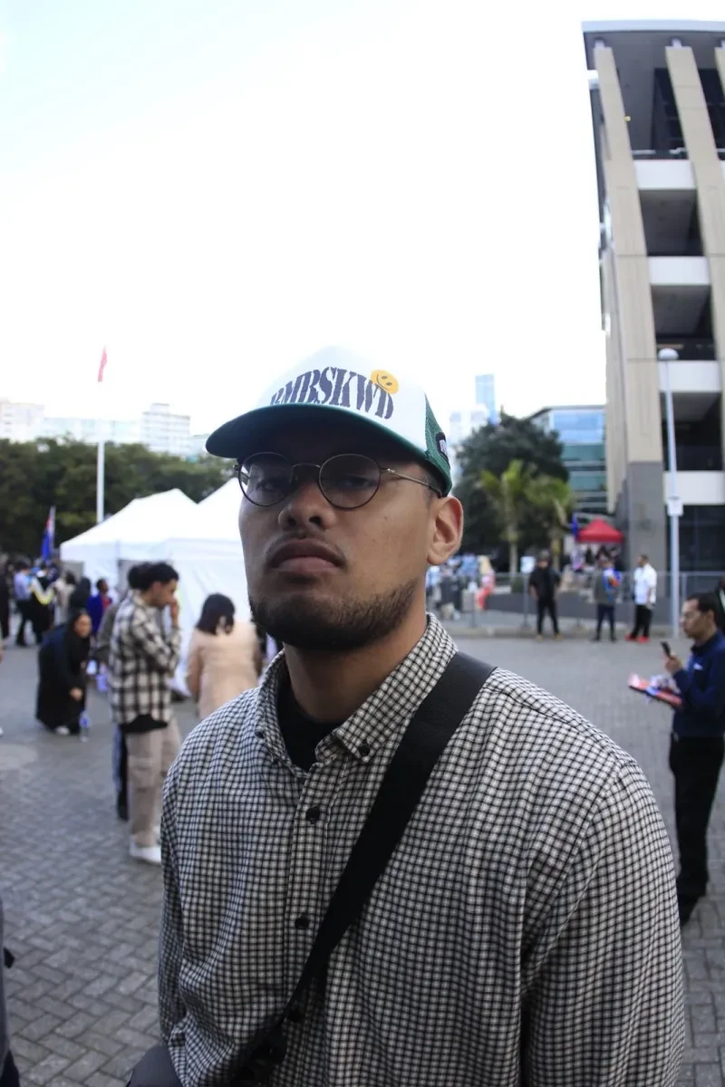 A young man wearing glasses and a cap with a short beard looks out of the picture. He's wearing a checked, button-down shirt and carries a black bag across his body