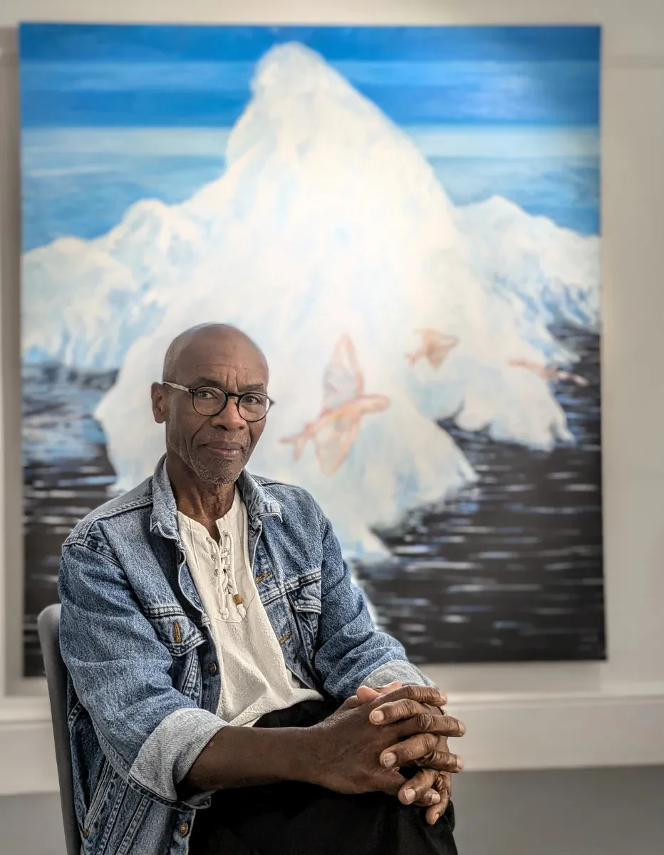 Artist Tam Joseph sits with his hands clasped across his knees. The painting 'Migrants' is in the background
