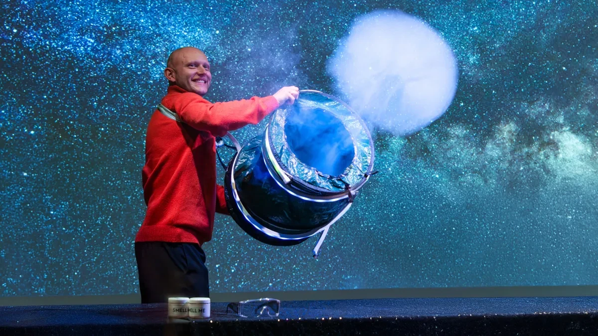 A smiling science explainer in a bright red jumper creates a cloud with a drum-like device on stage at Unboxing the Universe at the National Maritime Museum