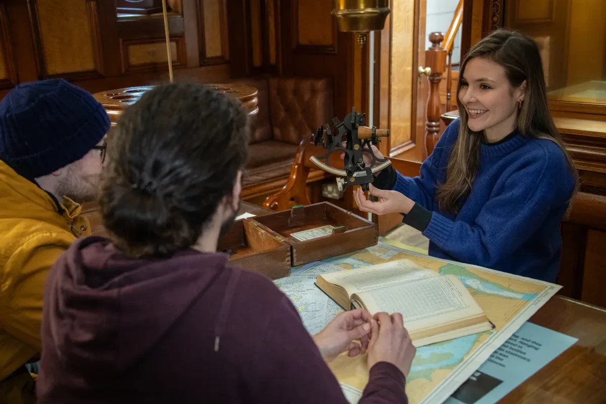 A visitor assistant sitting at captain's cabin on Cutty Sark gesturing towards an astronomical instrument in her hand
