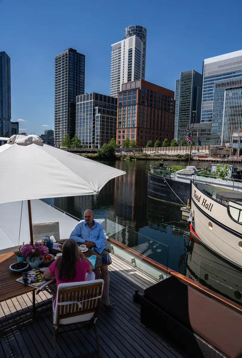 A couple on a boat sit underneath a parasol next to skyscrapers