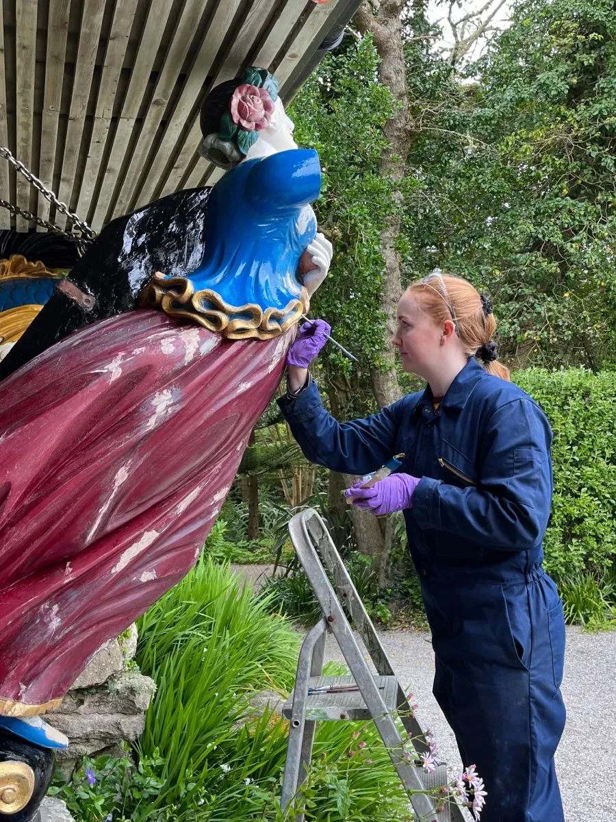 A woman in a blue boiler suit and purple gloves stands on a ladder and works on a red and blue painted figurehead statue in a garden