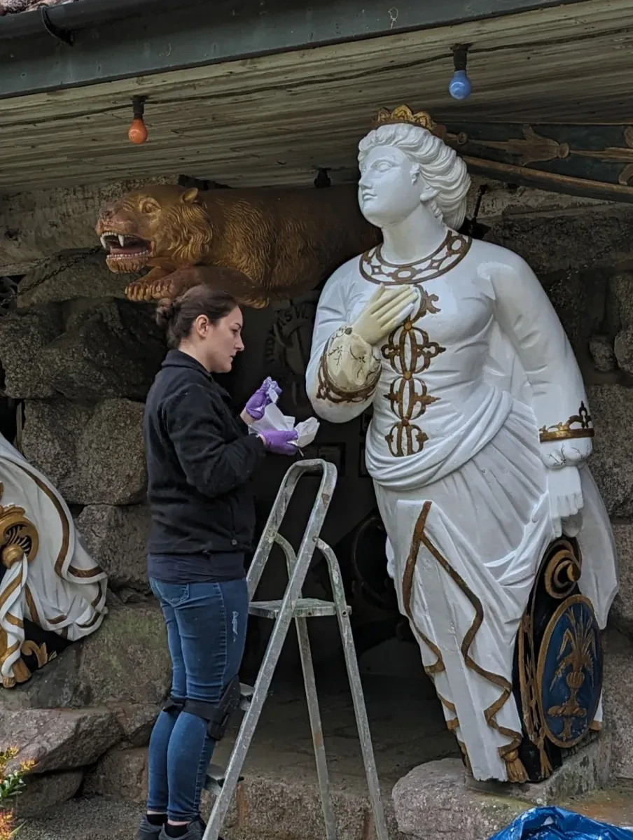 A woman in purple gloves stands on a ladder and repairs a classical white figurehead with an intricately carved face, hair, torso and dress