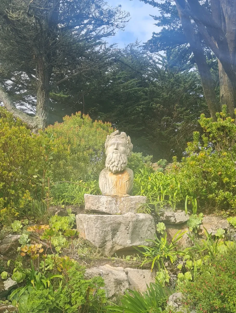 A carved, bearded figurehead stands in a garden surrounded by greenery
