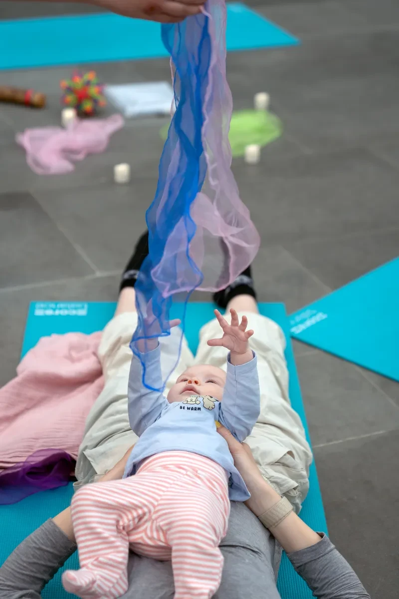 A baby lying on a parent's legs, reaching up to some fabric during a baby yoga session