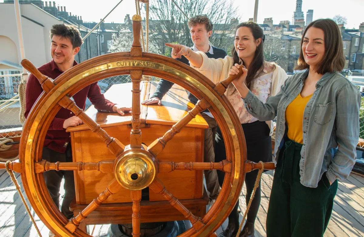Four smiling adults enjoy the view standing beside the replica ship's wheel on Cutty Sark