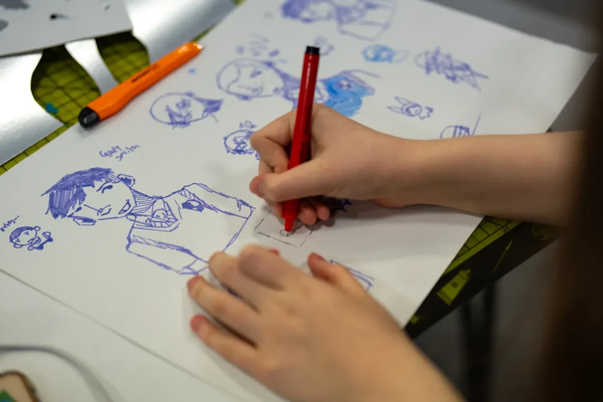 A young person is holding an ink pen and drawing cartoon people in their sketchbook