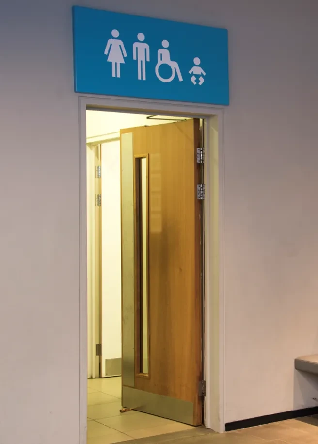 Photo of Romney Road entrance door into toilets with a blue sign above the door to show that there are male, female and access toilets within as well as baby changing