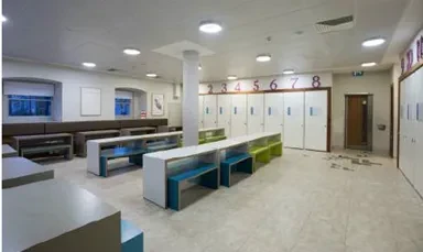 Photo of a classroom style room with long white tables, colour benches below, a column in the middle of the room, some large windows and cupboards around the outside.