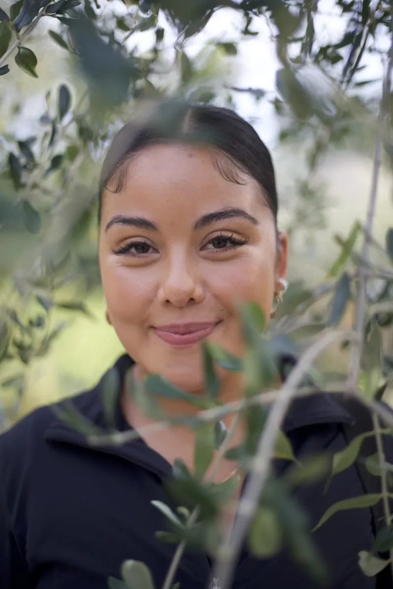 A headshot of Lirayen, looking through some branches. Lira is a London-based outdoor content creator, community builder, and nature enthusiast using her platform @OutsidewithLira to reframe what it means to belong in the outdoors.