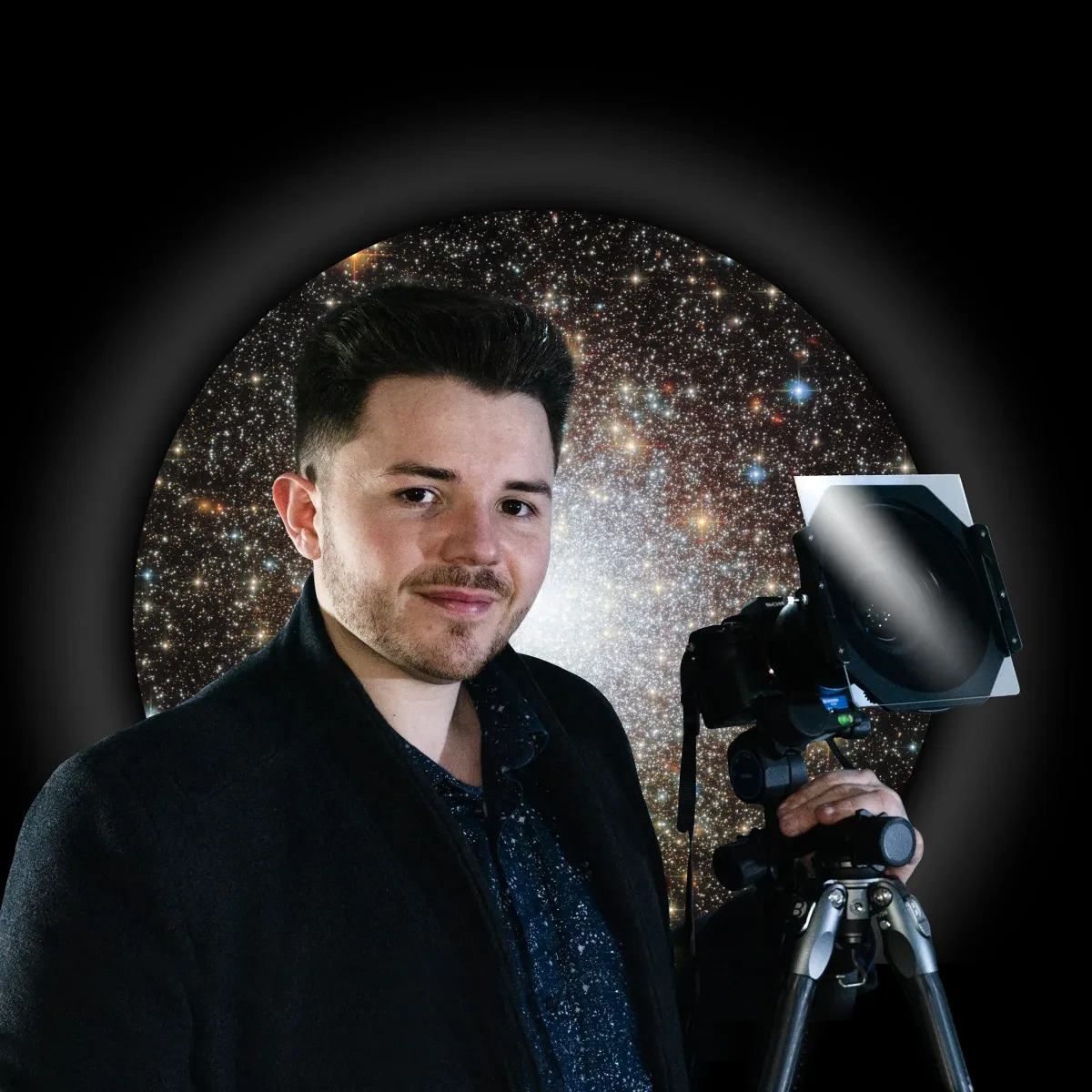 A young man with short dark hair stands facing the observer. He is wearing a dark suit jacket and blue shirt. Next to him is a camera and tripod set-up to look at the sky. In the background is a beautiful image of space containing thousands of beautifully coloured stars.